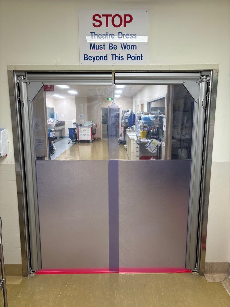 A double PVC swing door at a hospital operating theatre entrance: clear vision panels at eye height, grey lower panels and a red kick strip, in a stainless steel frame
